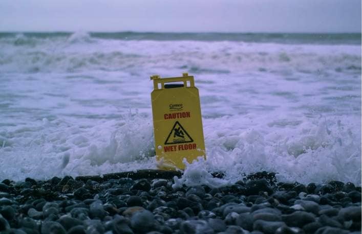 A Warning Wet Floor Sign Is Placed On The Beach And Is Covered By The Waves.  We Can Associate It With The Difficulties That Yiednodes Is Going Through In This Bearmarket.
